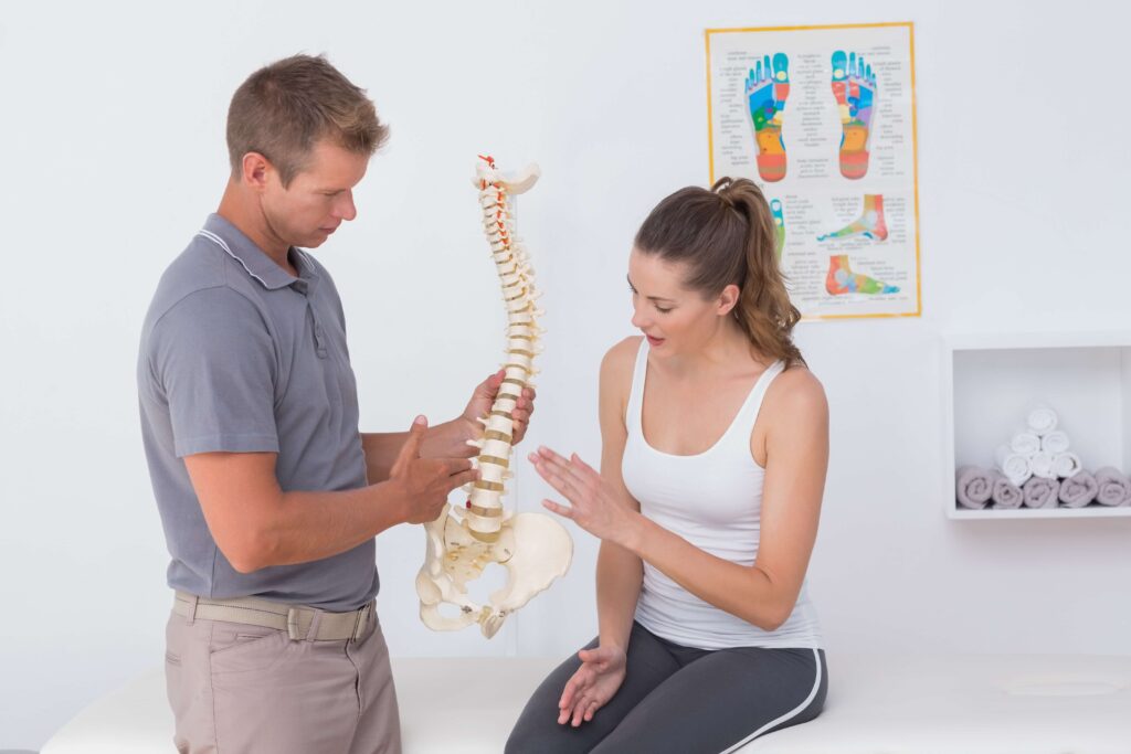 Female patient seated on clinic table being educated by male therapist using a model of a spine.
